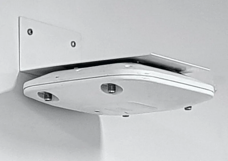 A white BAPLC bracket screwed to a pale wall, holding a white Cisco access point level underneath its arm, as if on a ceiling.