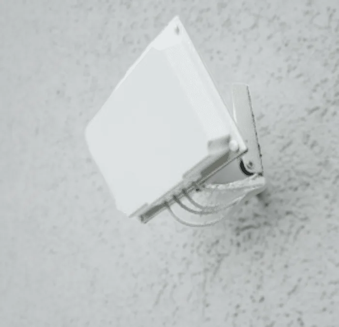 A flat white patch antenna on the hinged pan and tilt bracket, fixed to a textured rendered wall and angled down, with grey pigtail cables looped beneath it.