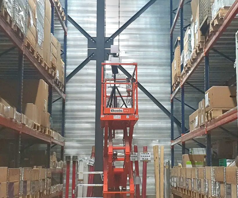 A Wi-Fi site survey in a high-bay warehouse: the survey access point on a tripod mast, raised on an orange scissor lift between tall pallet racking.