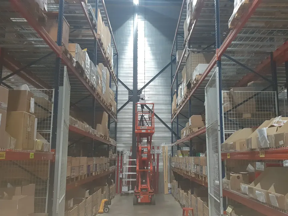 Survey kit on a mast at the top of an orange scissor lift, raised between tall pallet racking in a high-bay warehouse aisle.