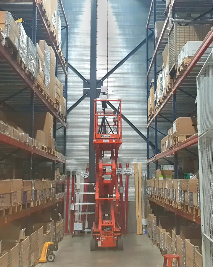 Close view of the survey kit on a mast at the top of an orange scissor lift, raised between tall pallet racking in a warehouse aisle.
