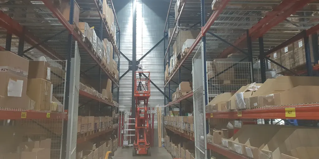 Survey kit on a short mast, raised on an orange scissor lift in a narrow aisle between high-bay racking stocked with cartons.
